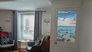 A modern living room with a view of a cityscape through a window, two grey armchairs, a small round table, and a door with a colorful stained glass design. - Harbour Tales (Weymouth)