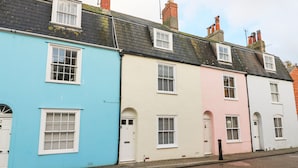 A row of three-story townhouses with distinct pastel colors, each featuring white-framed windows and doors, and a brick chimney. - Harbour Tales (Weymouth)
