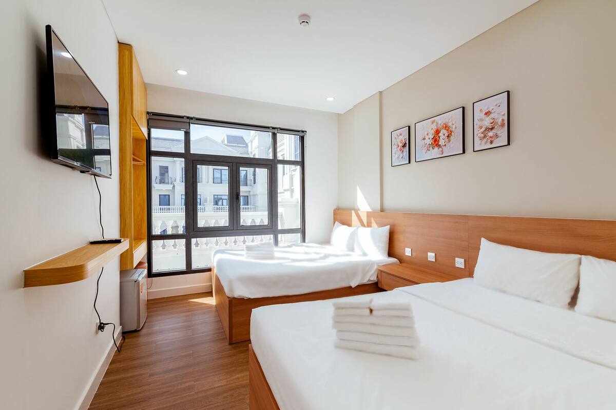 A hotel room with two beds, a TV mounted on the wall, and large windows offering a view of buildings outside.