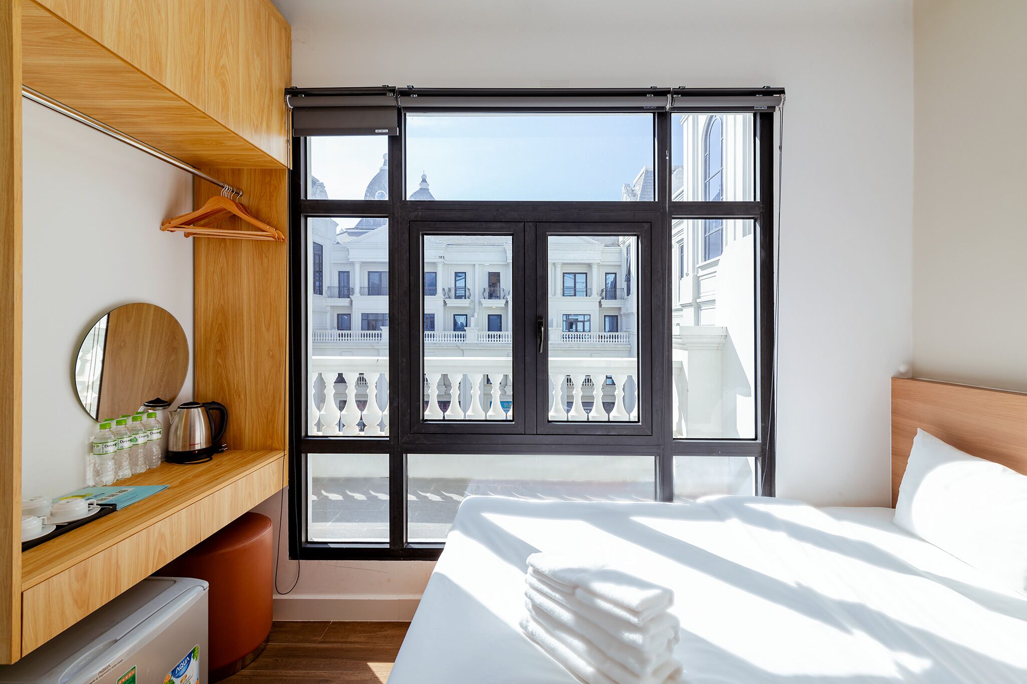 Quarto de hotel com cama, uma escrivaninha de madeira com espelho, cafeteira e vista para edifícios através de uma janela grande.