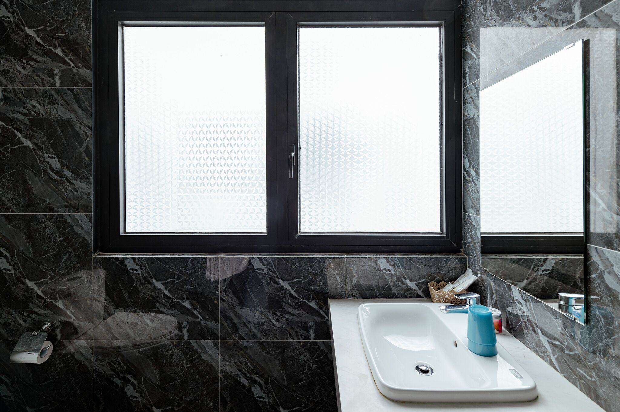 A bathroom with a large window, marble walls, a white sink, and a mirror.