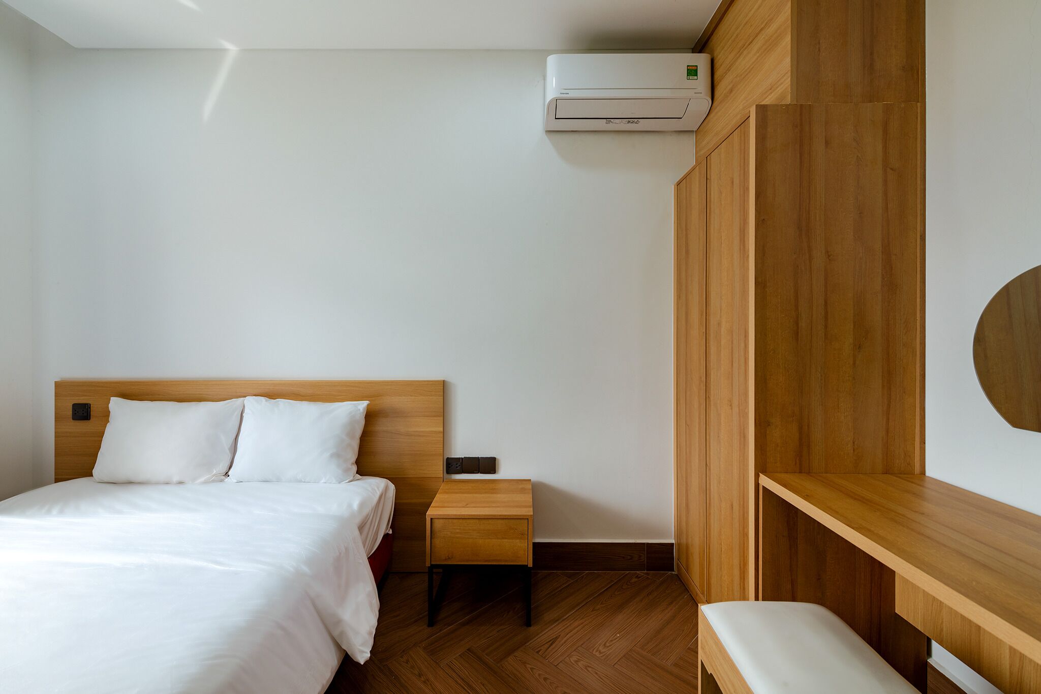 A hotel room with a wooden headboard bed, a bedside table, a bench, and a built-in wooden cabinet.