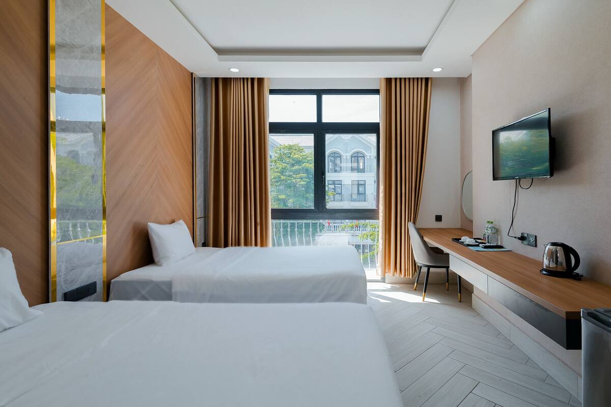 A hotel room with two beds, a desk, a TV, and a balcony with a view of a building.