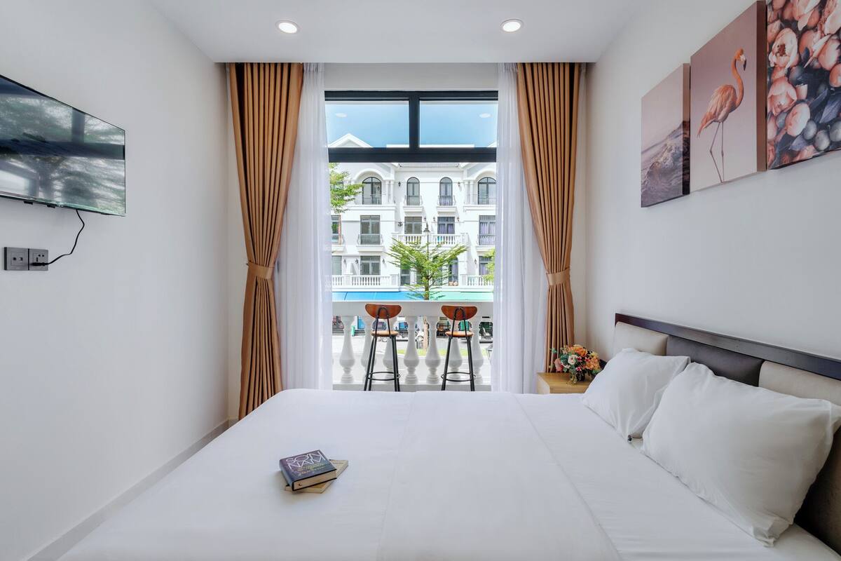 A hotel room with a large bed, a TV mounted on the wall, a balcony with a view of buildings, and a window with curtains.