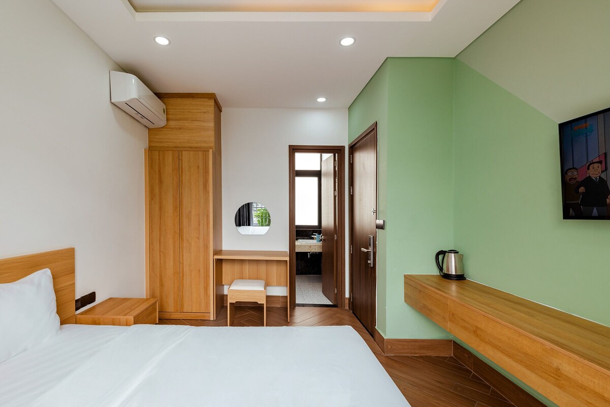 A hotel room with a bed, a wooden wardrobe, a small desk with a chair, a mirror, and a green wall.