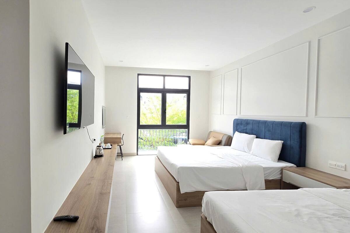 A modern hotel room with a large window, a flat-screen TV mounted on the wall, two beds, a wooden desk, and a chair.