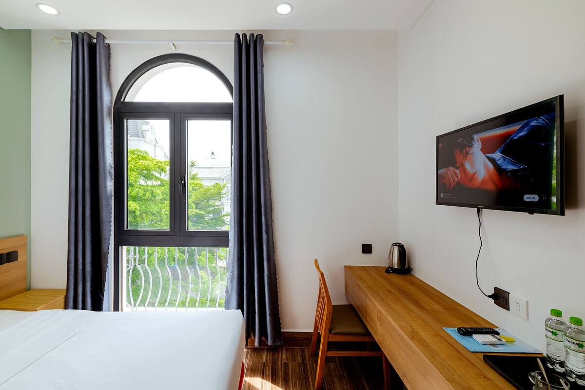 A hotel room with a bed, a wooden desk, a television, and a balcony with a view of greenery.