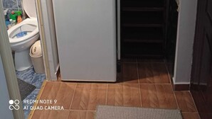Et kompakt køkken med et hvidt køleskab, et lille viktualierum og et toilet i baggrunden.