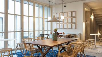 A dining area with a wooden table and wicker chairs, large windows, and framed artwork on the wall.