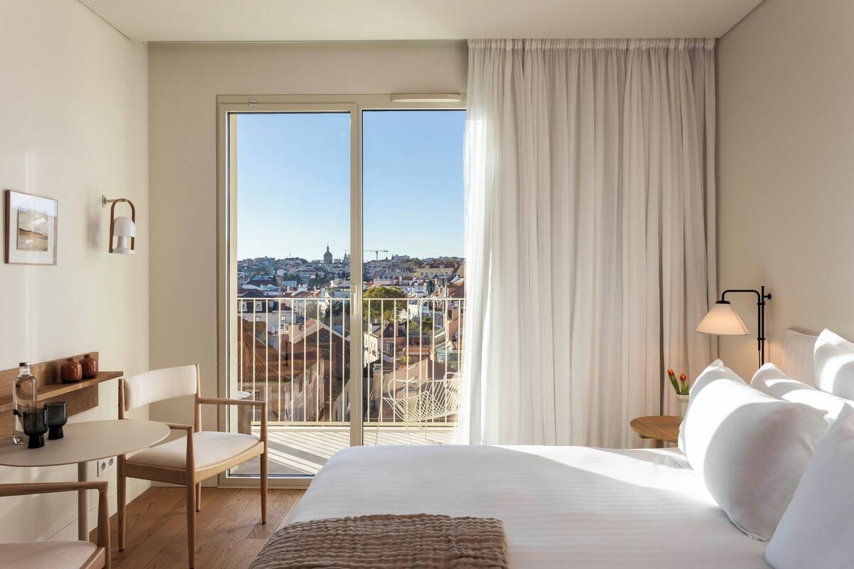 A hotel room with a large bed, a desk, and a chair. There is a view of a cityscape through a large window.