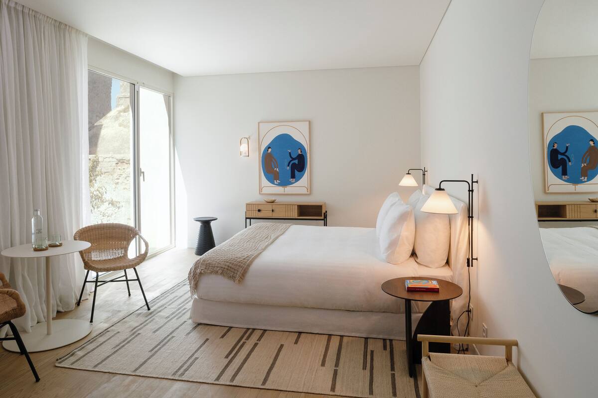 A modern hotel room with a large bed, a bedside table, a chair, a small table, and a window with curtains.