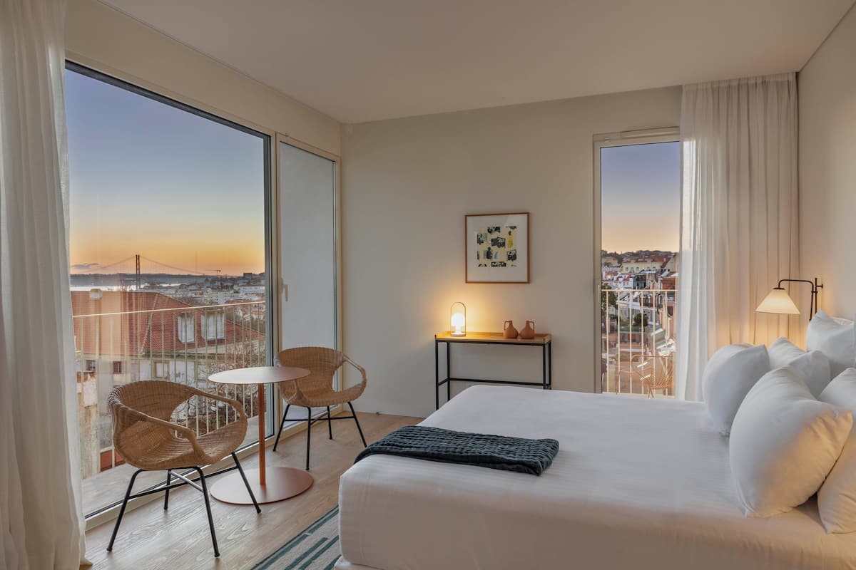 A hotel room with a large bed, two wicker chairs, a small table with a lamp, and a city view through large windows.