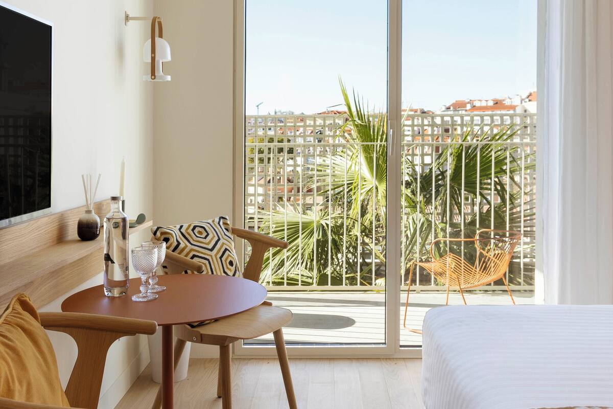 A hotel room with a bed, a TV, a dining table with chairs, and a balcony with a view of palm trees and buildings.