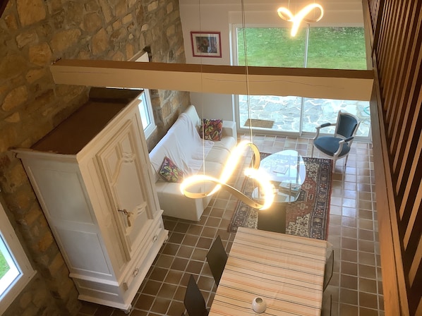 A cozy room with a stone wall, a white cabinet, a sofa with colorful cushions, a glass coffee table, and a view of the outdoors through large windows.