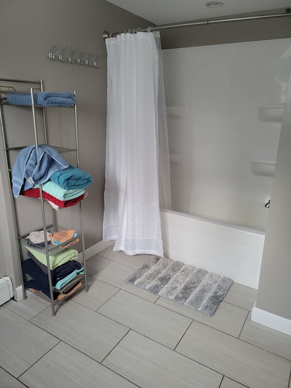 A bathroom with a shower, a metal towel rack with folded towels, and a grey rug.