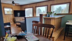A cozy cabin interior with wooden walls, a dining table with a notebook and pen, a chair, and a television in the background.