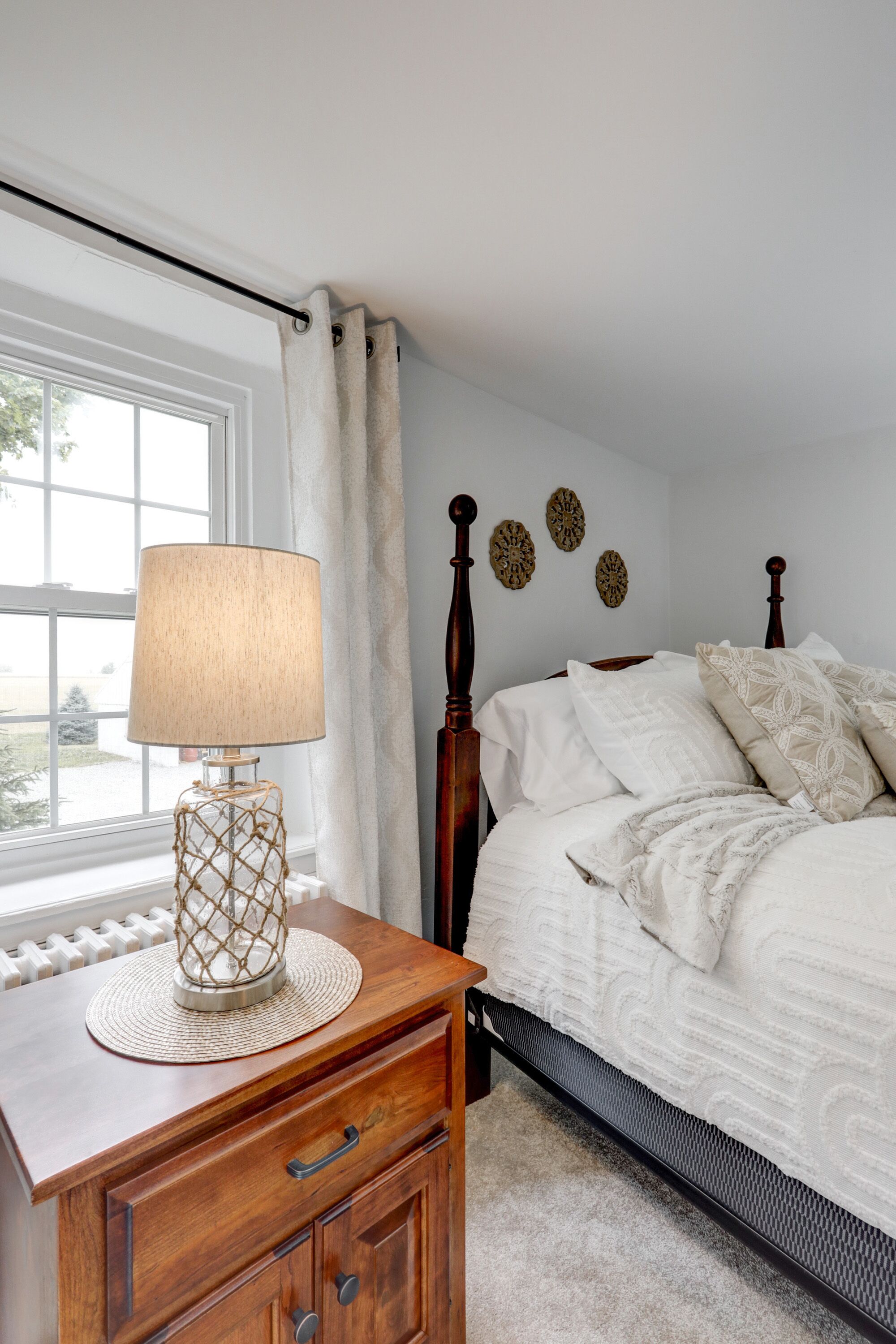 A bedroom with a wooden nightstand, a lamp, a bed with white bedding, and a view of the outdoors through a window.