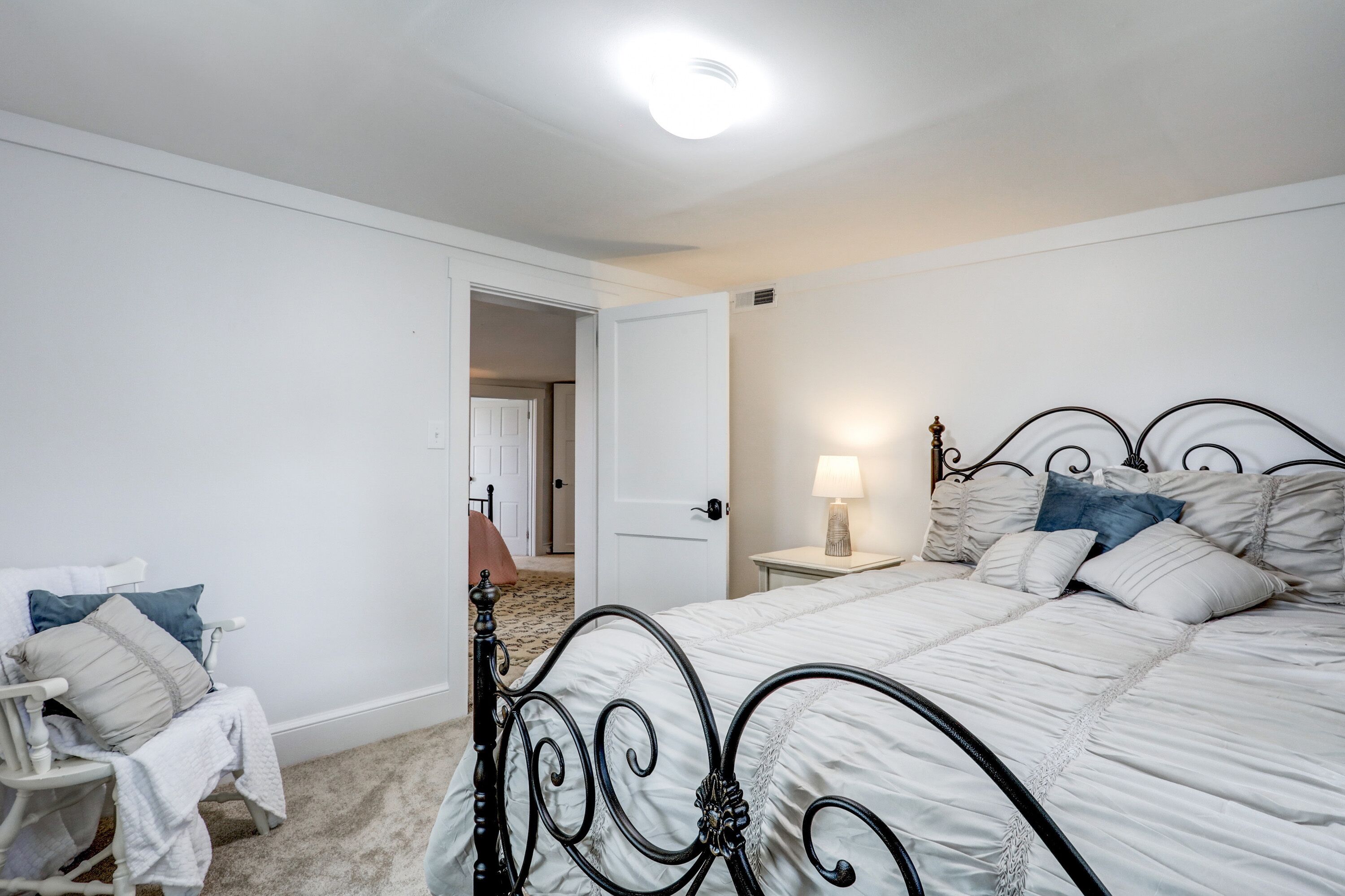 A bedroom with a bed, bedside table, lamp, chair, and an open door leading to another room.