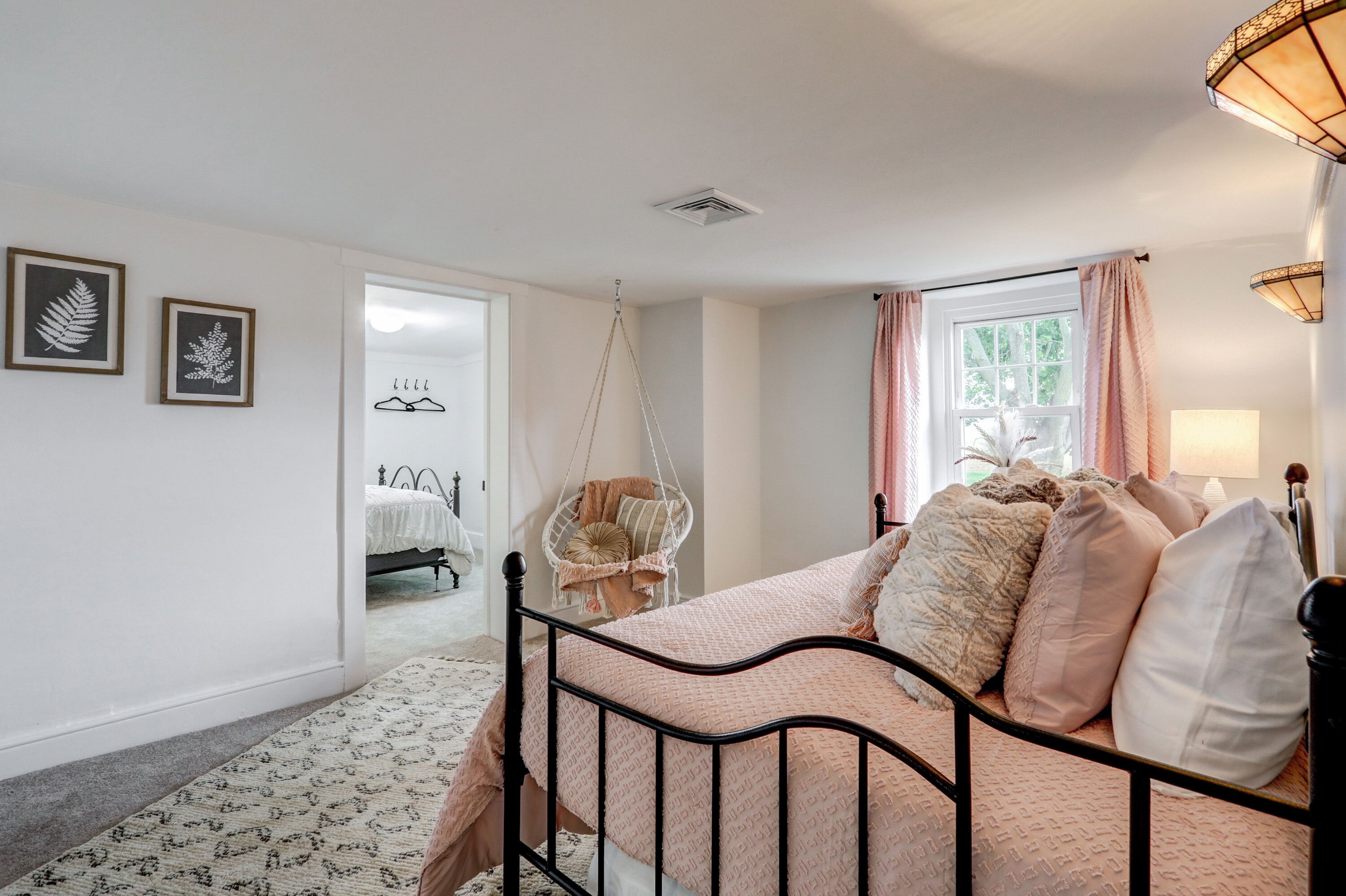 Un dormitorio con cama, una silla colgante, una ventana con cortinas y una puerta que da a otra habitación.