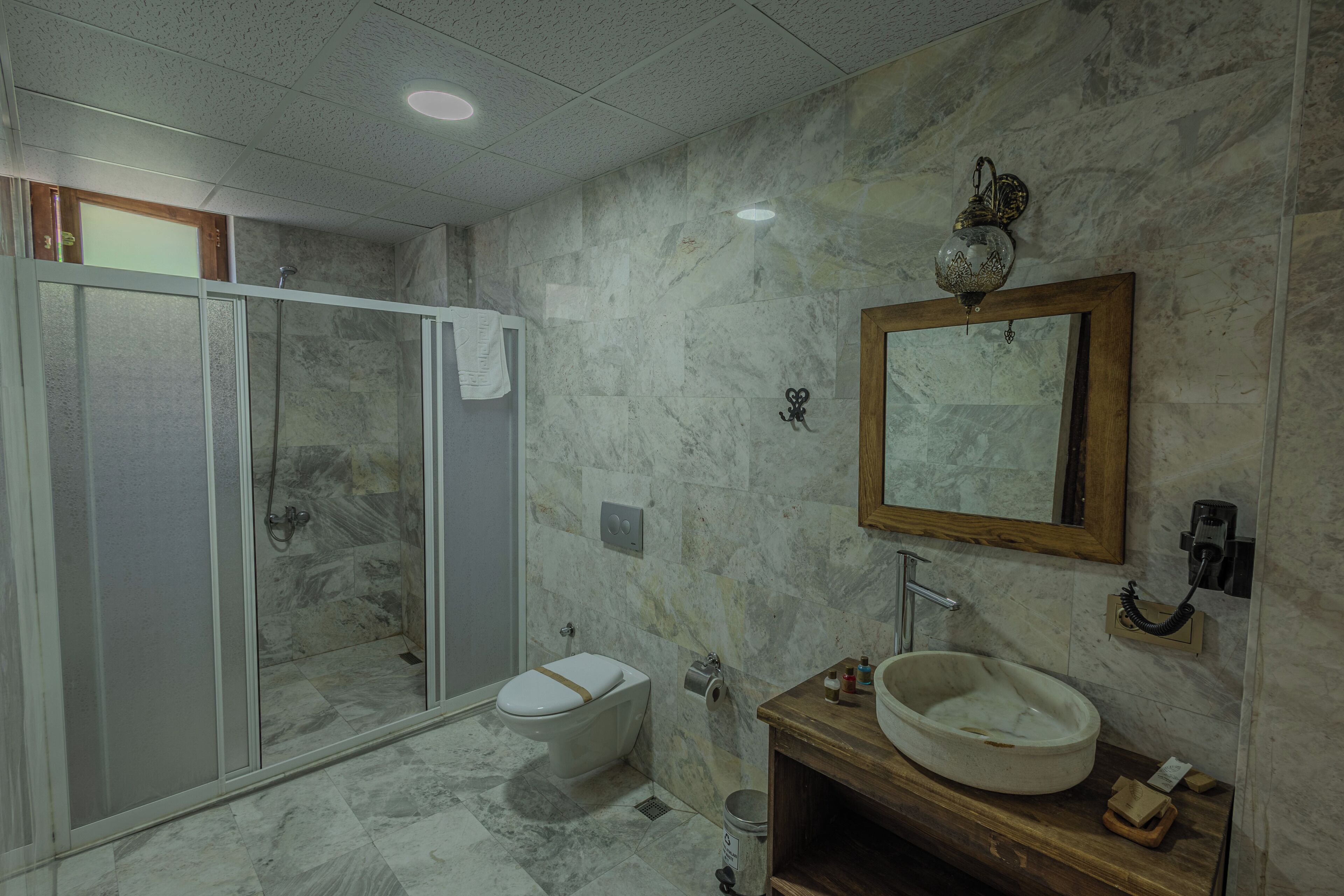 Ein Badezimmer mit einer Glaskabine zur Dusche, einer Toilette, einer Wand mit Marmorplatten, einem Holzkosmetikschrank mit Waschbecken und einem Spiegel.