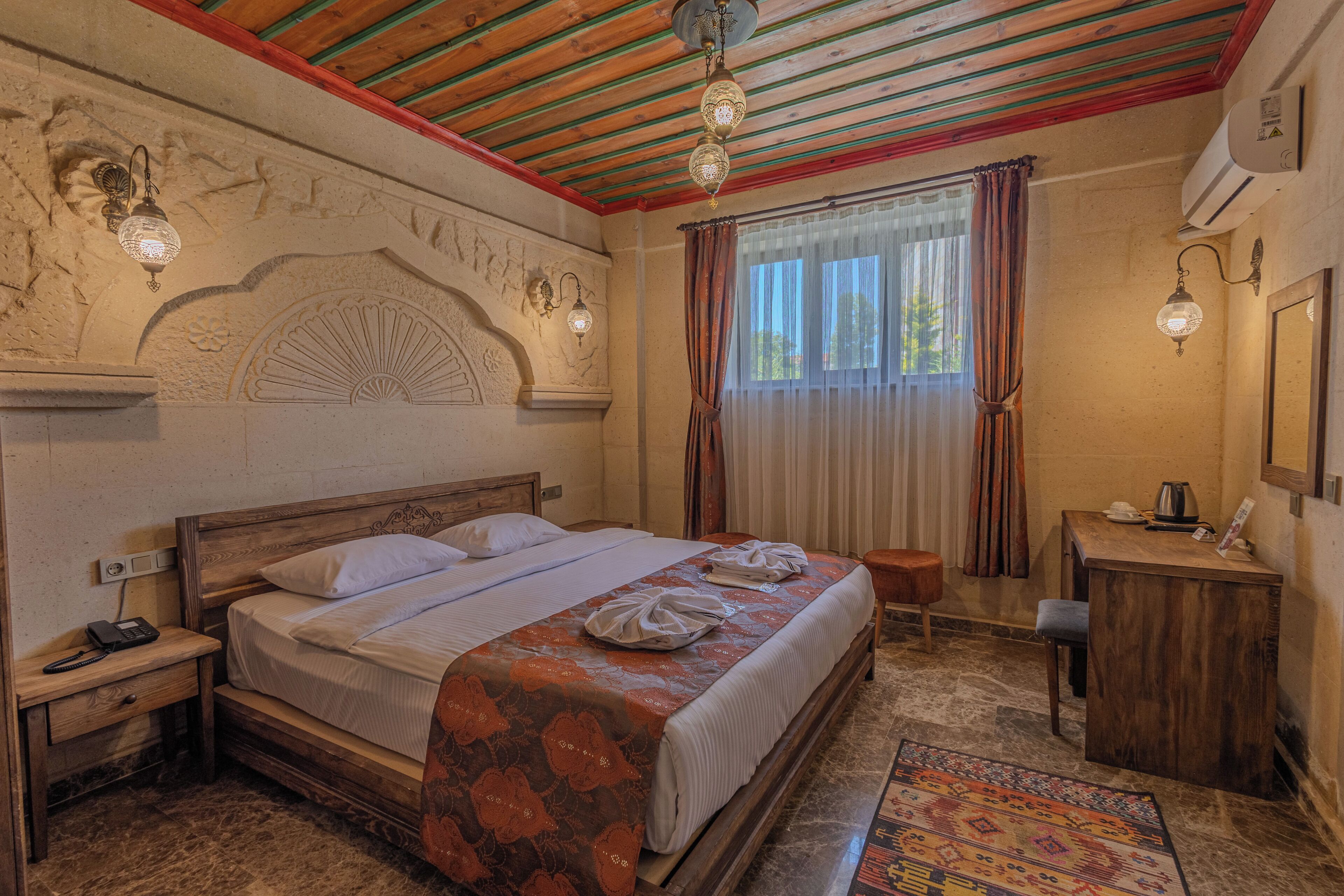 A bedroom with a wooden bed, a nightstand with a phone, a desk with a kettle, and a patterned rug.