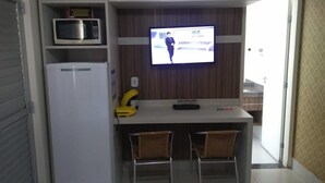 Cocina compacta equipada con microondas, refrigerador y un televisor que muestra un noticiero. Hay dos taburetes de bar y una mesita pequeña.
