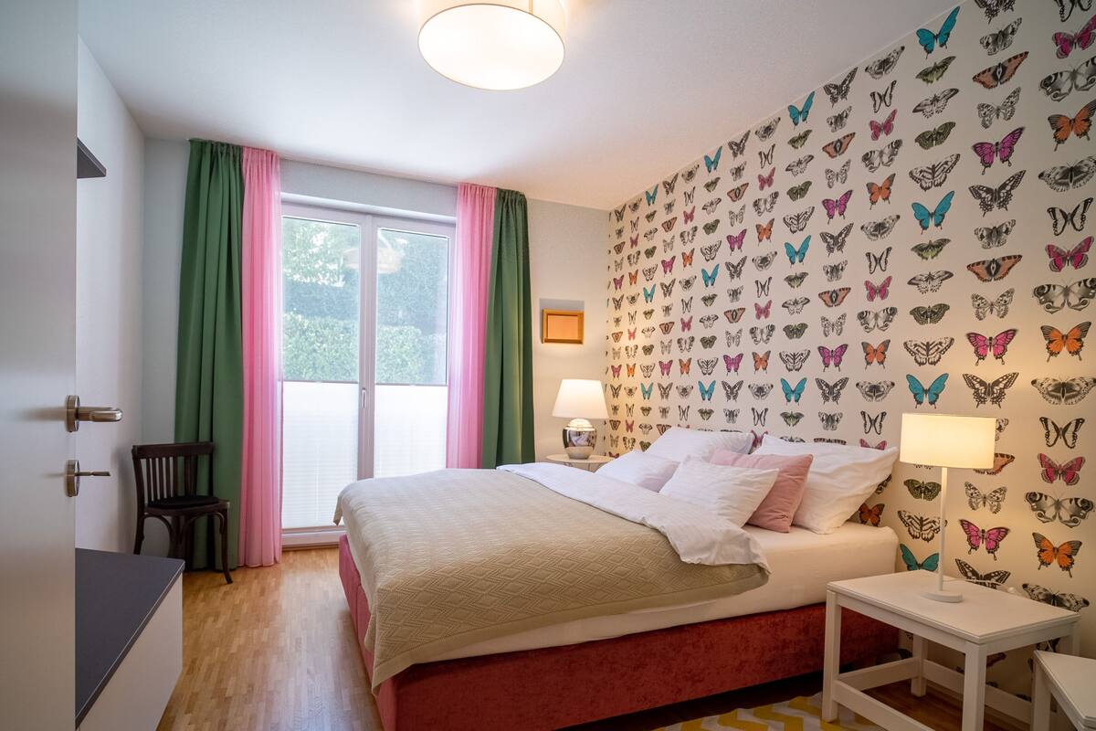 A bedroom with a bed, a nightstand, a chair, a window with curtains, and a wall with butterfly decals.
