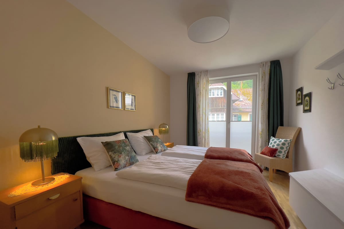 A bedroom with a bed, bedside tables, a lamp, a chair, and a view of the outside.