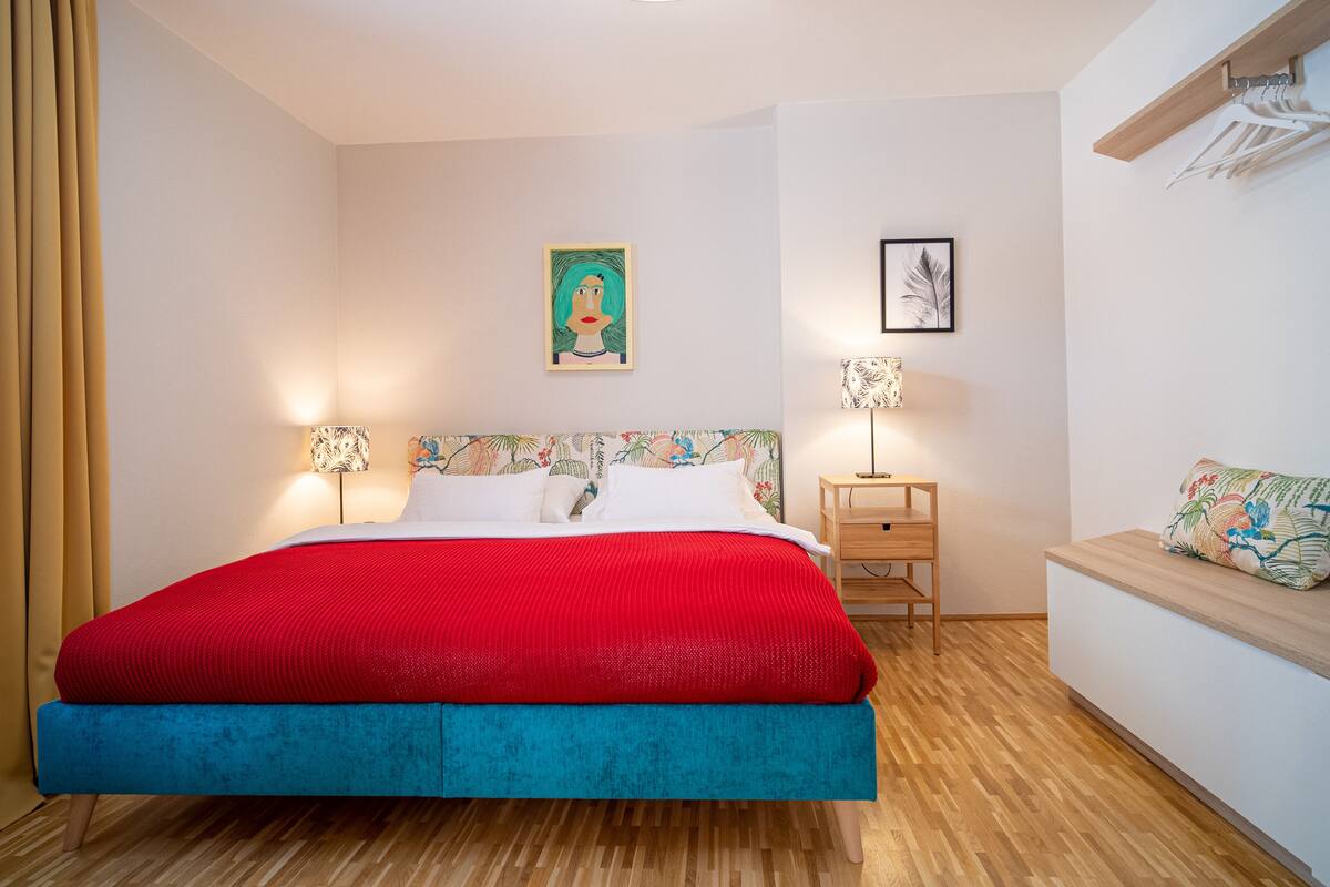 A bedroom with a bed, bedside tables, a nightstand, a wall-mounted shelf, and a hanging light fixture.