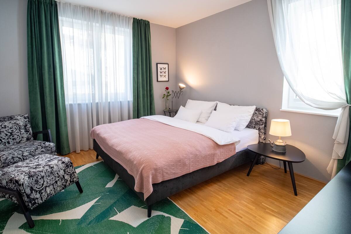 A neatly arranged bedroom with a bed, a chair, a small table with a lamp, and a wall-mounted picture.