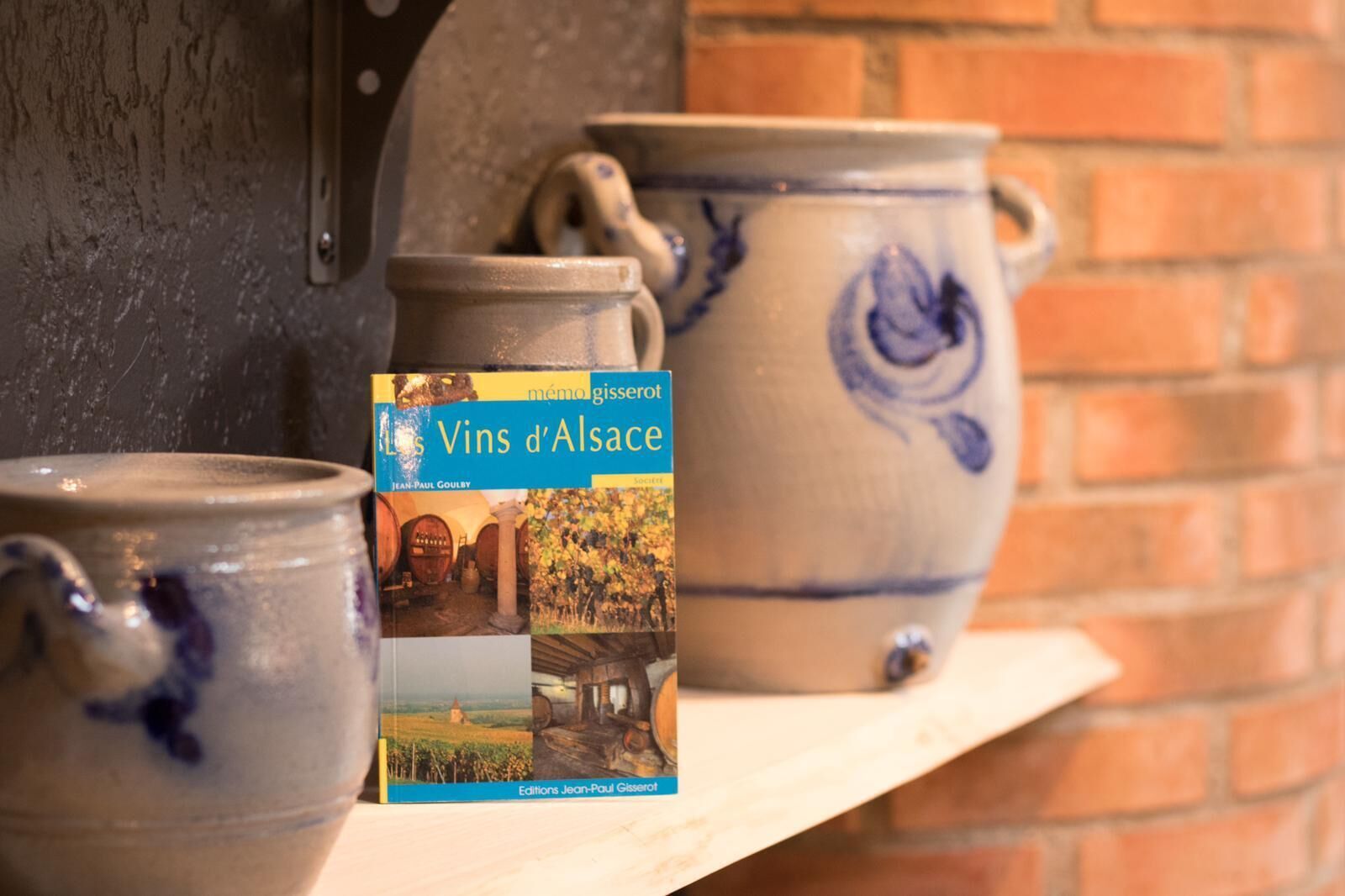 Um livro intitulado 'Les Vins d'Alsace' de Jean-Paul Gisserot, colocado entre vasos de cerâmica numa estante.
