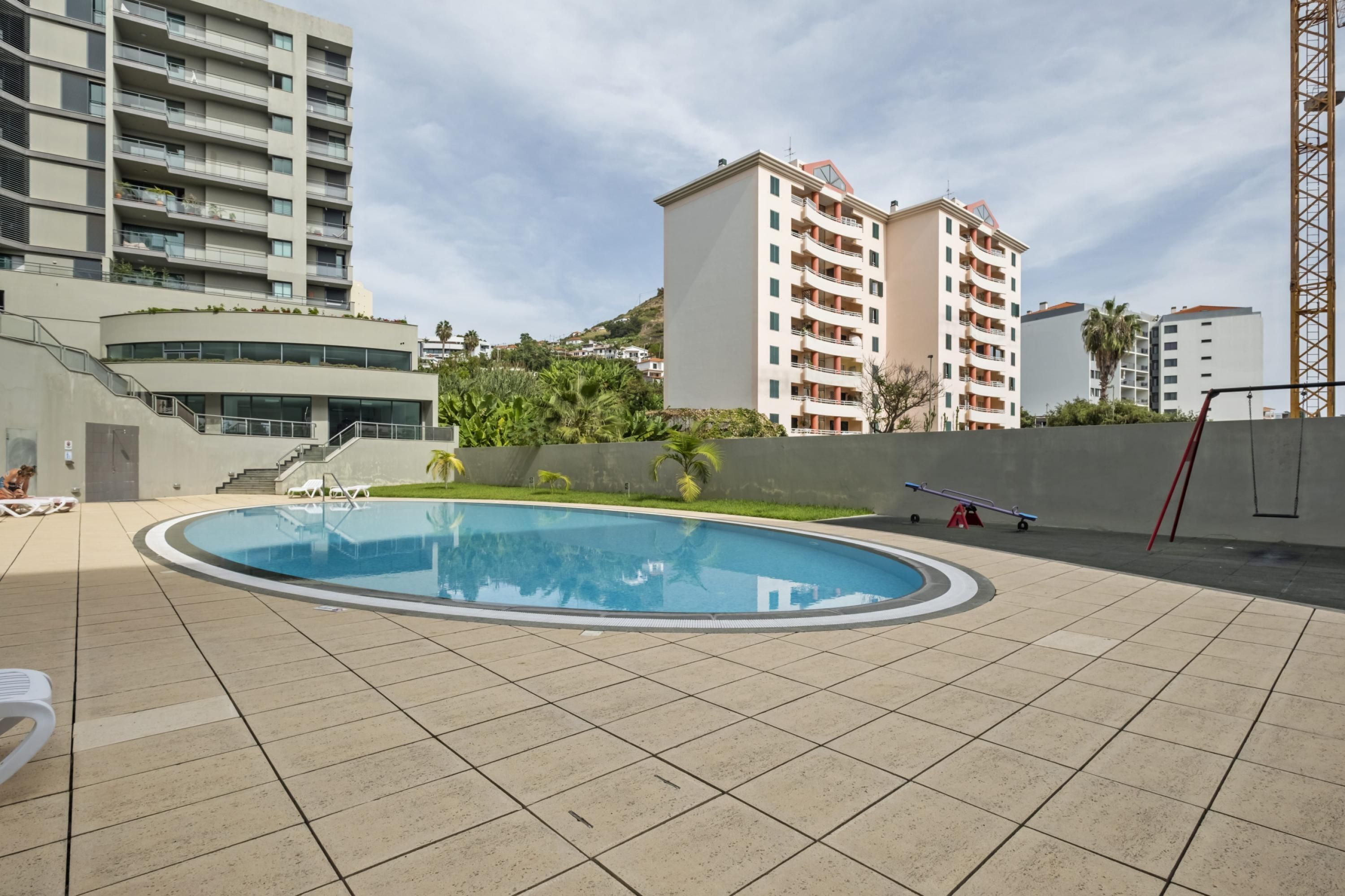 Una piscina con un borde de baldosas, rodeada de edificios y una colina al fondo.
