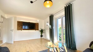A modern living room with a glass coffee table, dark sofas, a wooden accent wall, and a view of greenery outside. - Modern vacation apartment in the Allgäu (Erkheim)