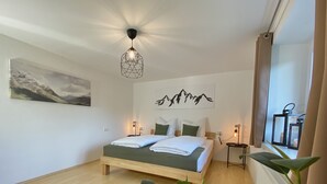 A modern bedroom with a wooden bed, a geometric pendant light, a mountain art piece, and a view of the outdoors. - Modern vacation apartment in the Allgäu (Erkheim)