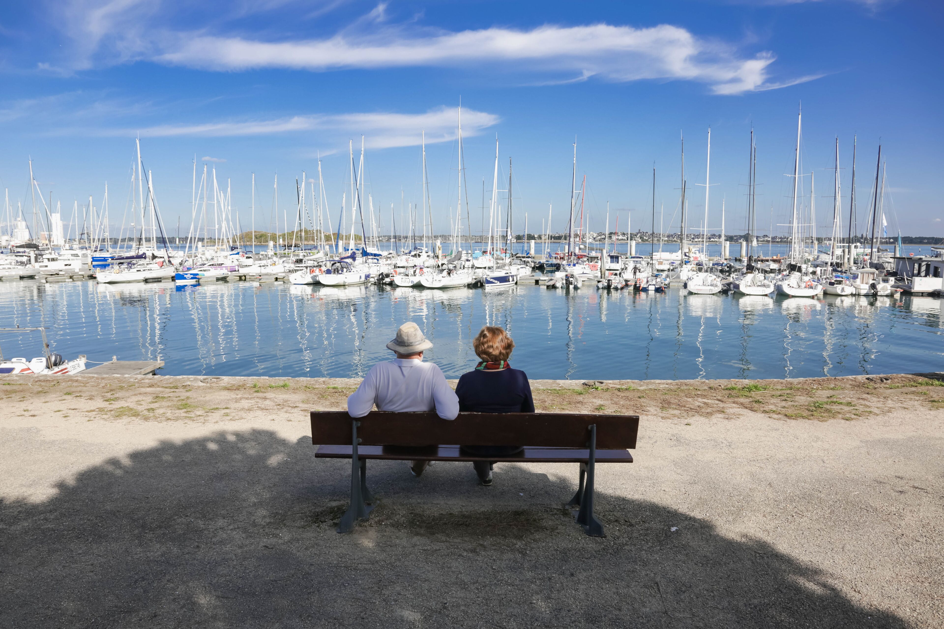 A couple sitting on a bench overlooking a marina with numerous sailboats.