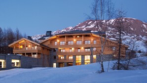 A large, multi-story wooden building with balconies and large windows, surrounded by snow and mountains. - Lagrange Vacances L'Alpenrose (Huez)