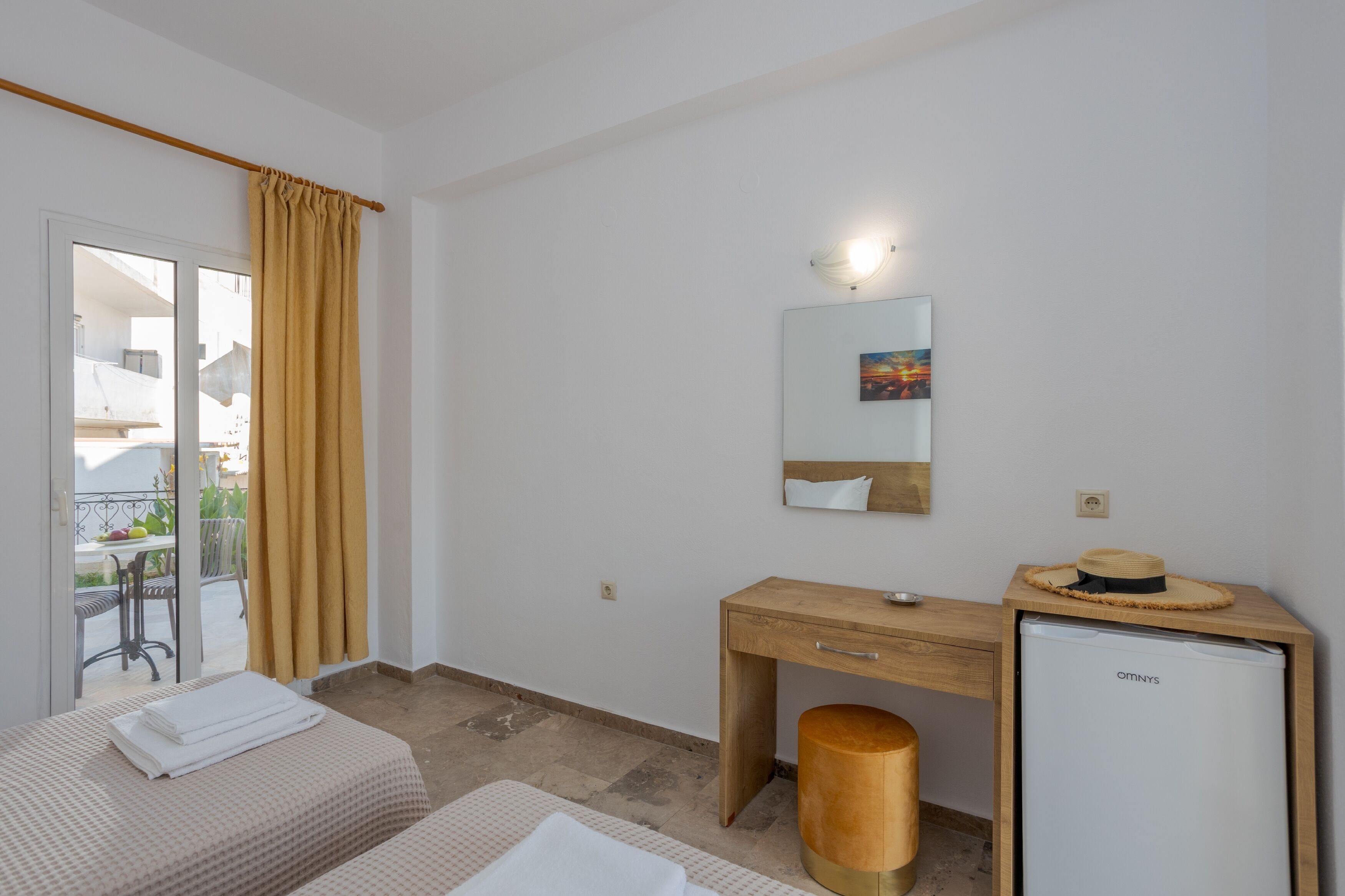 A hotel room with a bed, a wooden desk, a small fridge, a mirror, and a balcony with yellow curtains.