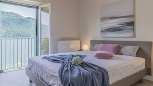 A bedroom with a bed, a bedside table, a nightstand, a wall-mounted lamp, and a view of a balcony with a mountain and water. - Sunset View Lierna_varennaholidays (Lierna)