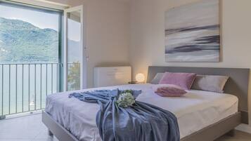 Camera da letto con un letto, un comodino, una lampada a parete e vista su un balcone con montagna e acqua.