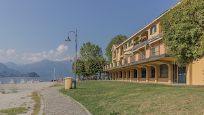 A coastal building with a paved walkway, a grassy area, and a view of the sea and mountains in the background. - Sunset View Lierna_varennaholidays (Lierna)