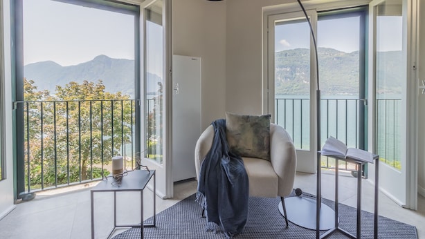 A modern living room with a sofa, a small table, and a standing lamp. The room has large glass doors leading to a balcony with a view of mountains and a body of water. - Sunset View Lierna_varennaholidays (Lierna)