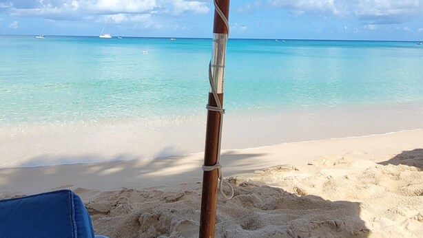 A beach umbrella with a wooden frame and a blue canopy, positioned over a sandy area with a clear view of the ocean and distant boats. - Beautiful  4BR villa w/pool in St James, Porters.  5 min walk to gorgeous beach. (Porters)
