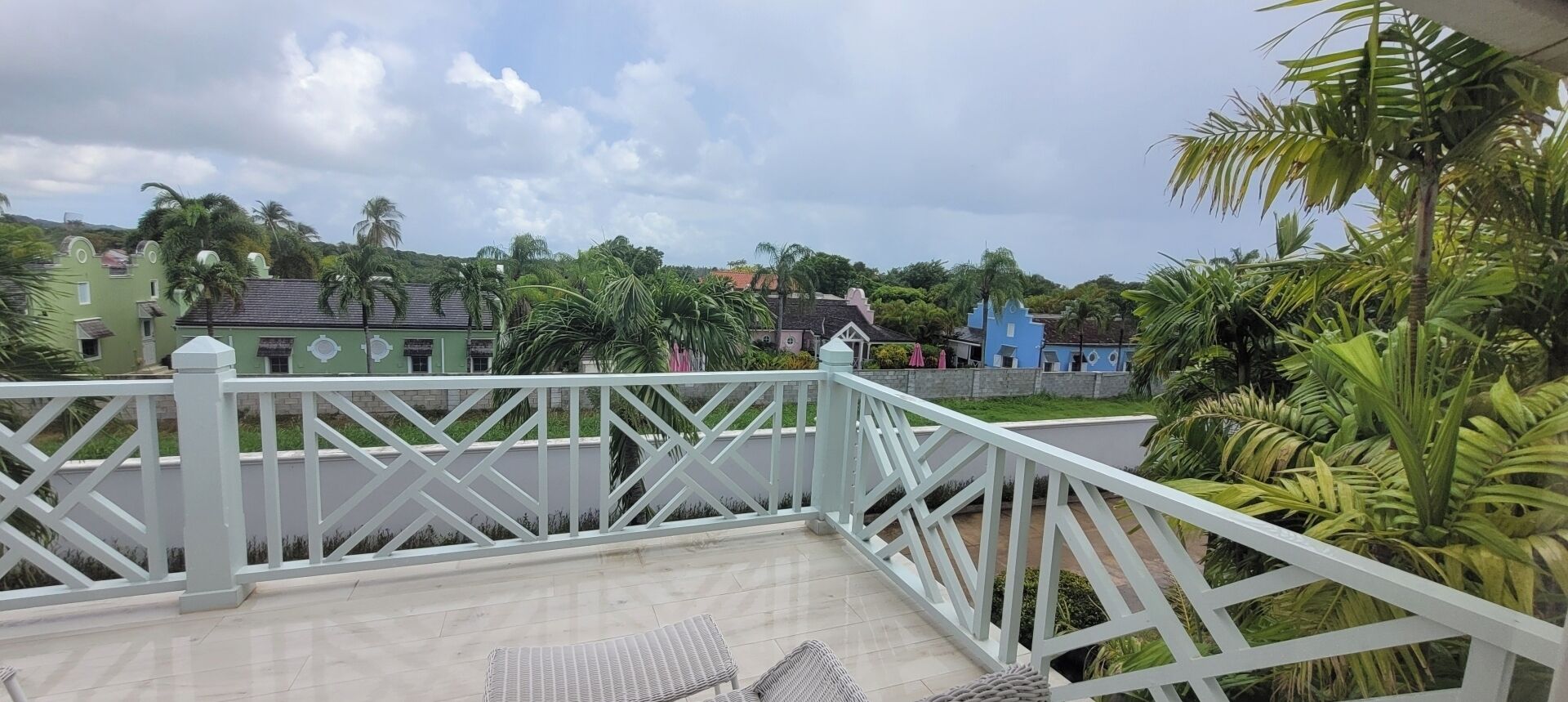 Un balcón con barandilla blanca, suelo de baldosas y vista a una zona residencial con casas y palmeras.