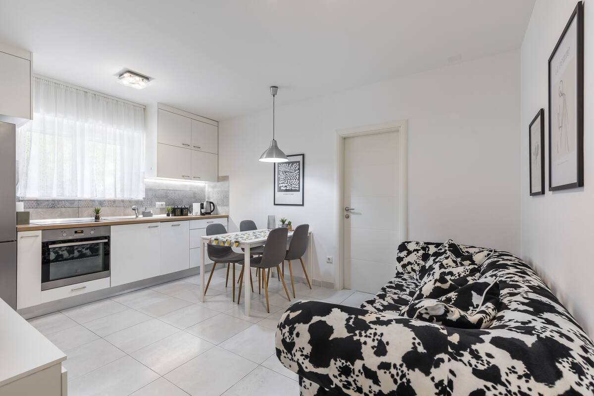 A modern kitchen with white cabinets, a dining table with chairs, and a black and white patterned sofa in a living area.