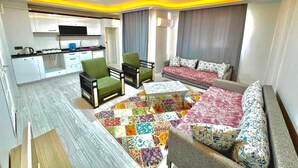 Un salon moderne avec un canapé, des fauteuils, une table basse et un tapis coloré. On y trouve également un coin cuisine avec des meubles blancs et une cuisinière à gaz.