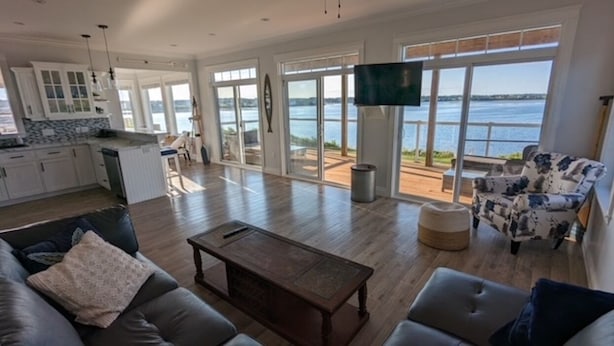 Ein geräumiger Wohnbereich mit Holzboden, einer Couch, einem Couchtisch und einem Fernseher. Ein großes Fenster bietet Blick auf das Wasser.
