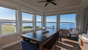Ein Zimmer mit einem großen Holztisch, einem blauen Stuhl und Blick auf ein Gewässer durch große Fenster.