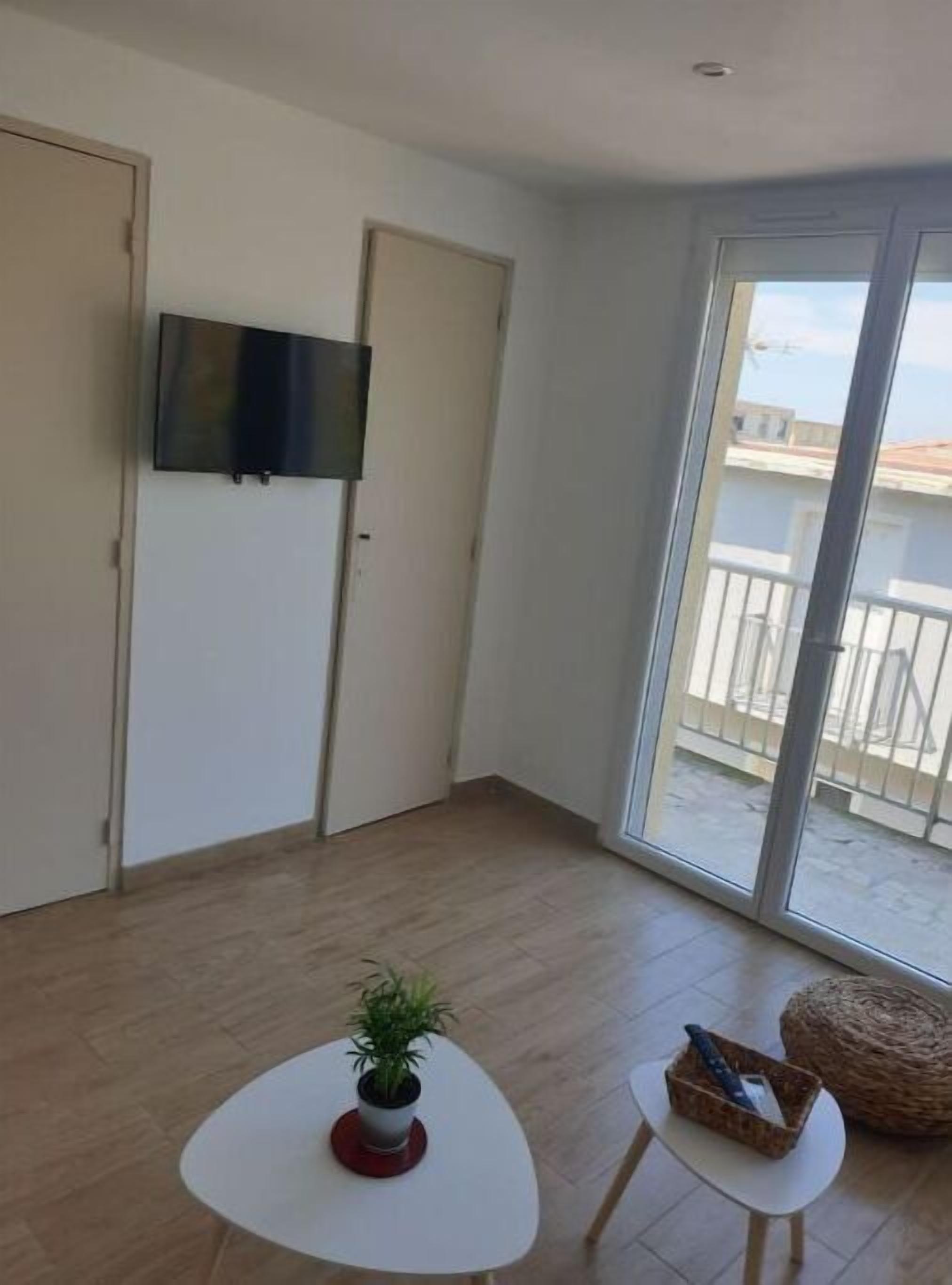 Ein Zimmer mit einem an der Wand befestigten Flachbildfernseher, einer Glastür zu einem Balkon und einem kleinen, runden Tisch mit einem Blumentopf.
