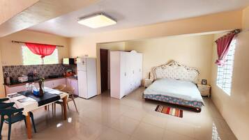 A room with a bed, a desk with chairs, a kitchen area, and a wardrobe.