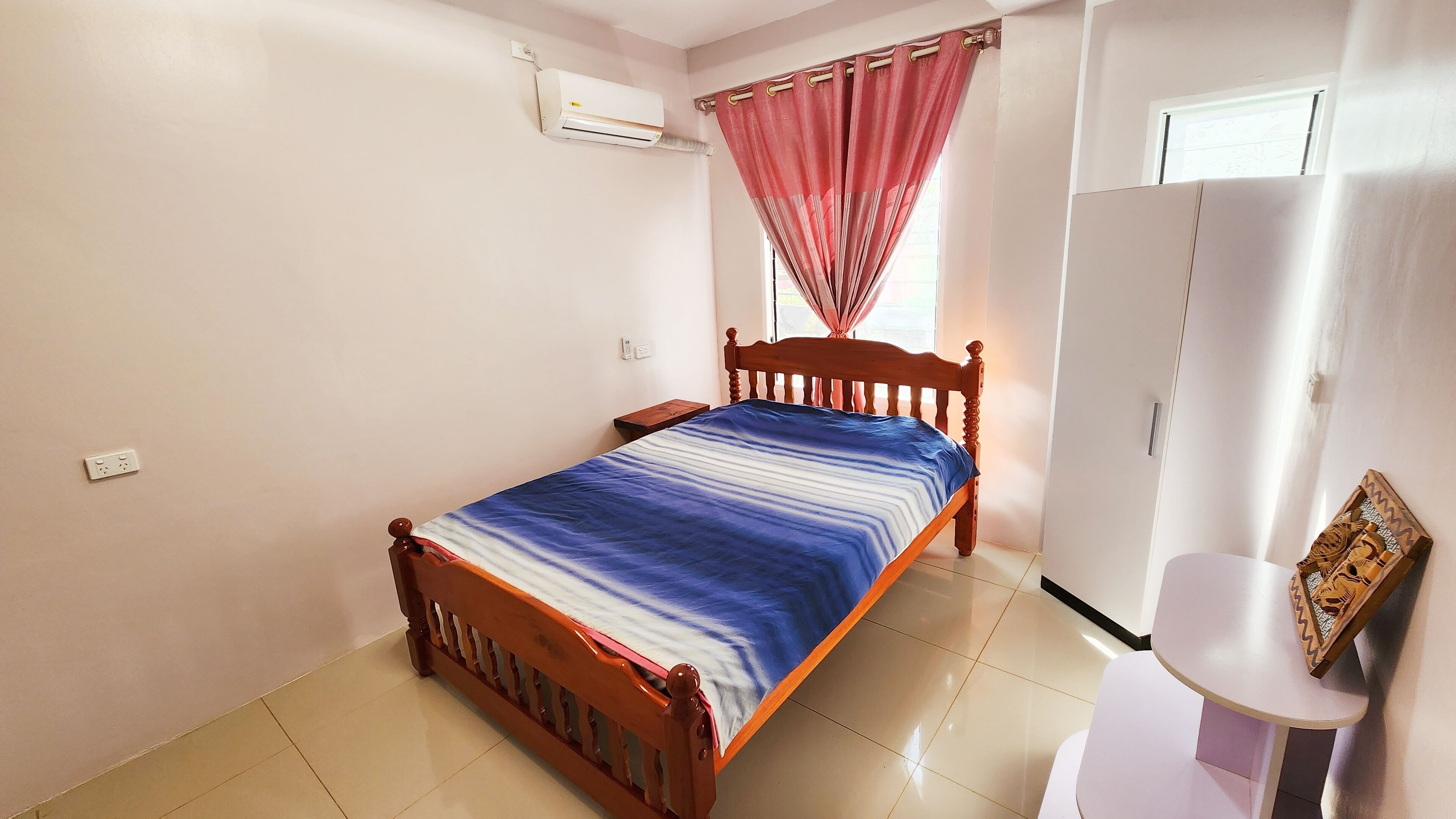 A bedroom with a wooden bed, a white wardrobe, a small table, and a window with curtains.